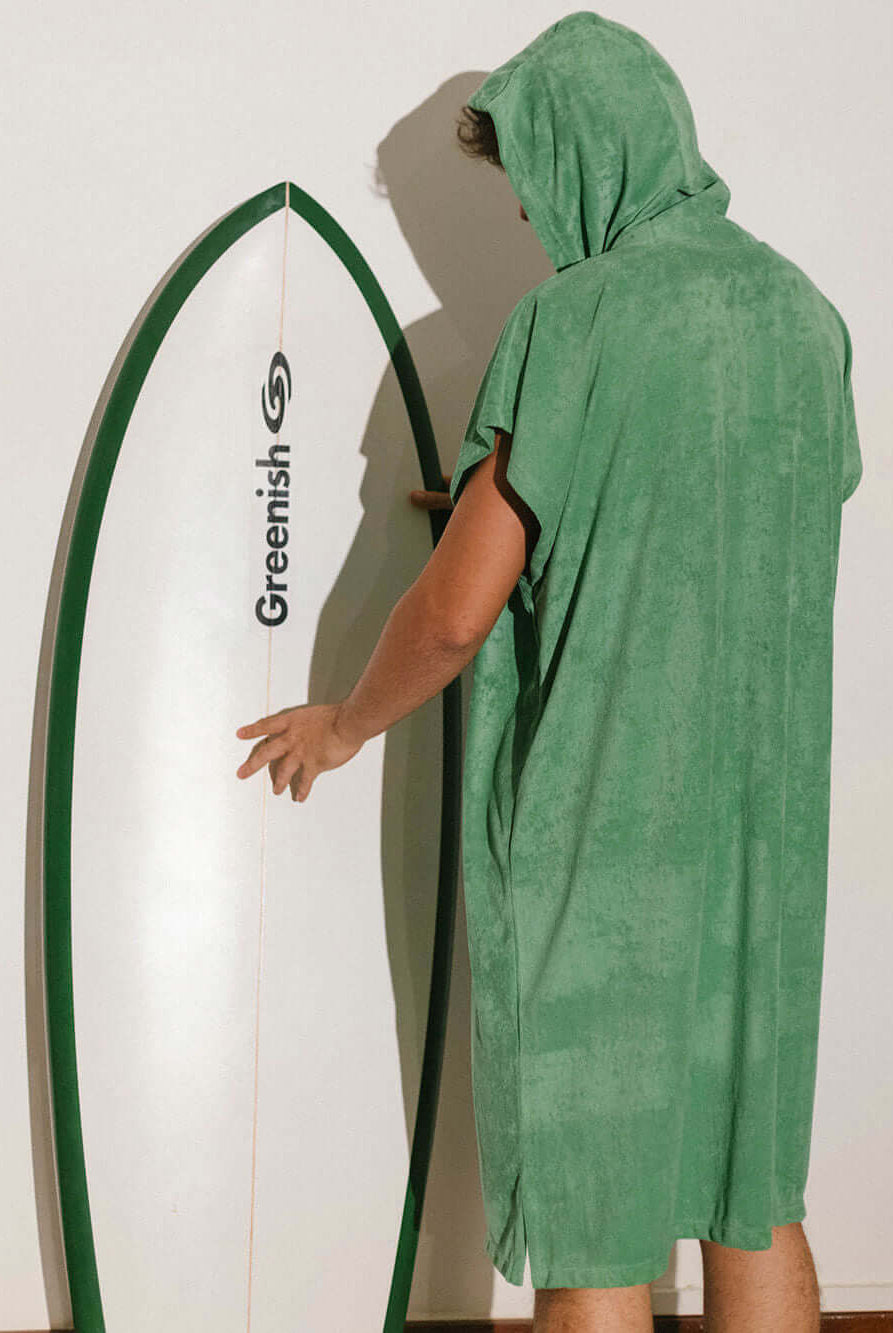 Homem usando poncho adulto Greenish verde ao lado de uma prancha de surf, ideal para praia e esportes aquáticos.