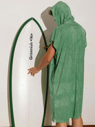 Homem usando poncho adulto Greenish verde ao lado de uma prancha de surf, ideal para praia e esportes aquáticos.