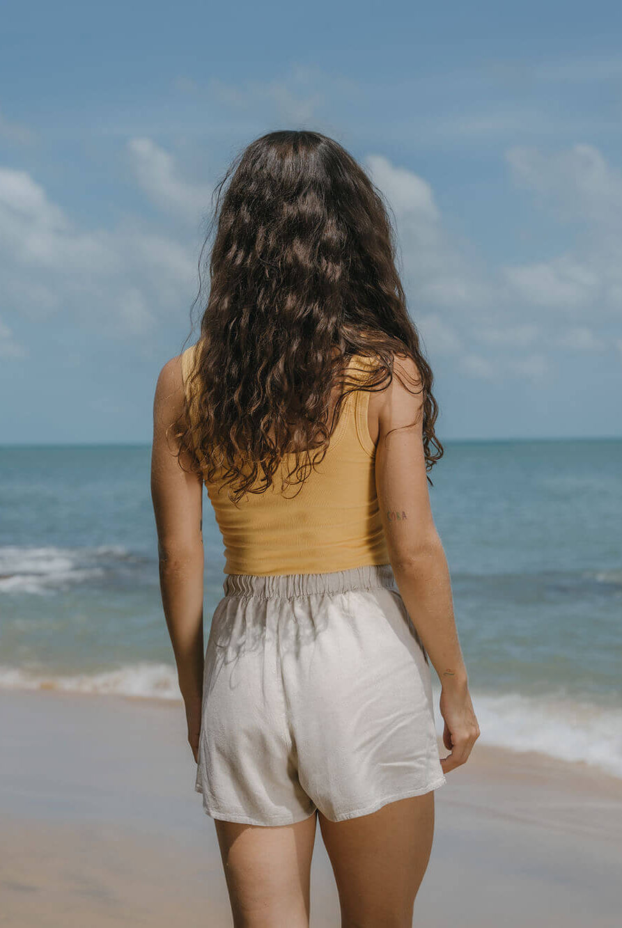 Mulher de costas na praia usando regata Kora amarelo da Greenish, refletindo estilo e conforto.