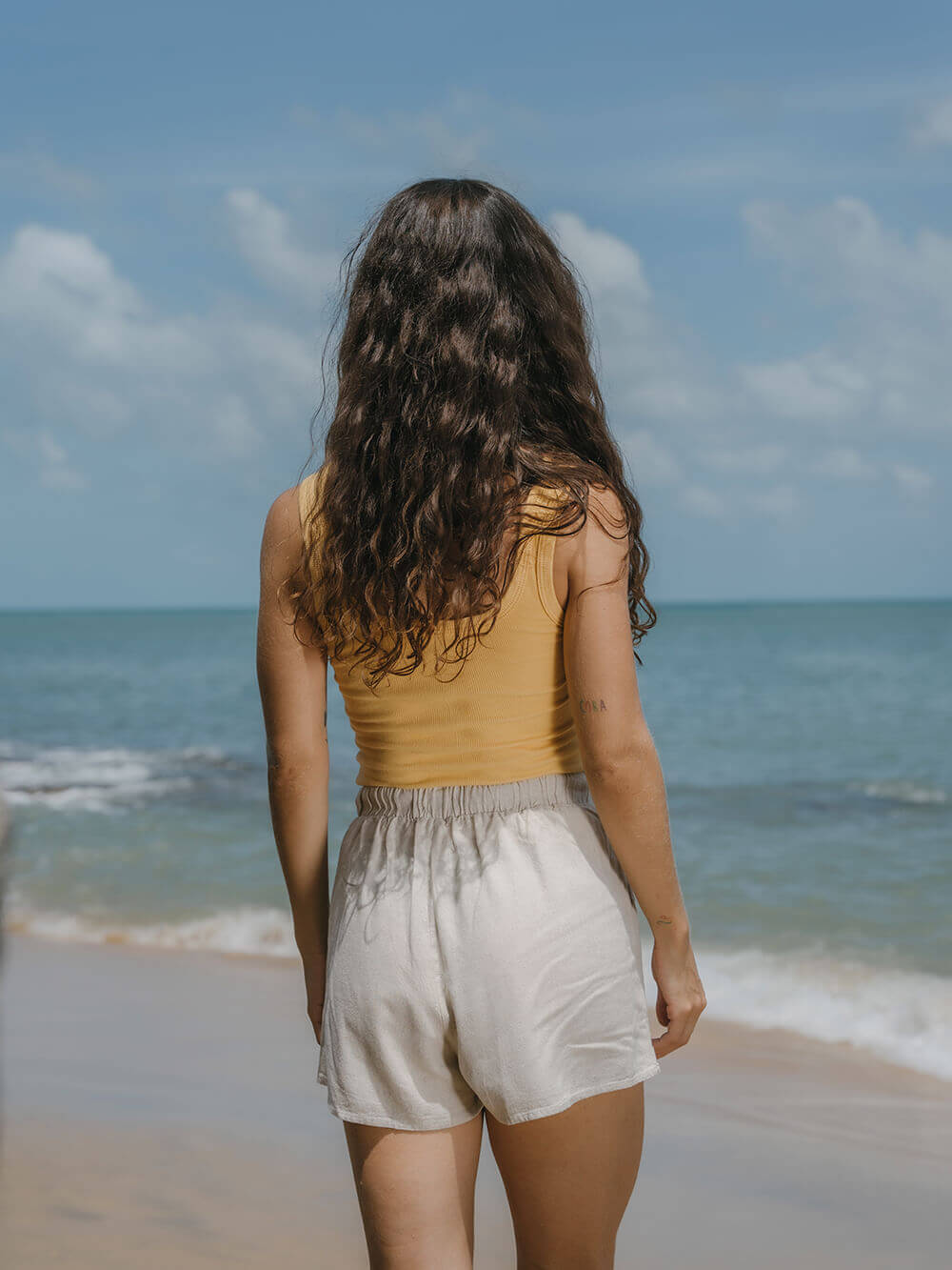 Mulher de costas na praia usando regata Kora amarelo da Greenish, refletindo estilo e conforto.