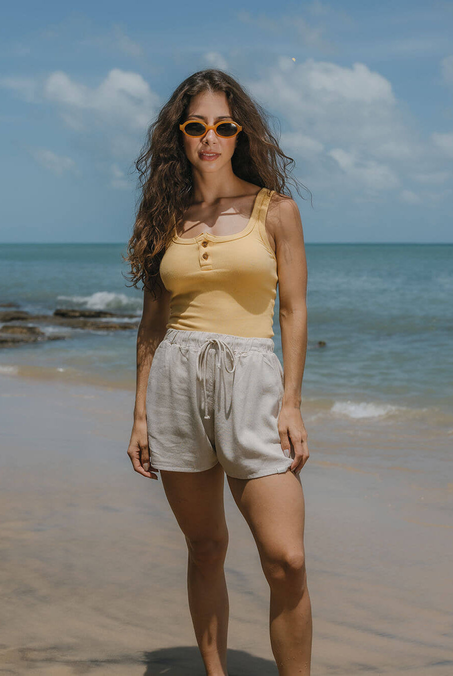 Modelo usando regata Kora amarela da Greenish na praia, com shorts claros e óculos de sol, ideal para dias quentes.