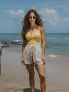 Modelo usando regata Kora amarela da Greenish na praia, com shorts claros e óculos de sol, ideal para dias quentes.