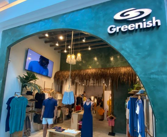 Fachada da loja Greenish com cabides de roupas e decoração tropical, destacando estilo e modernidade.