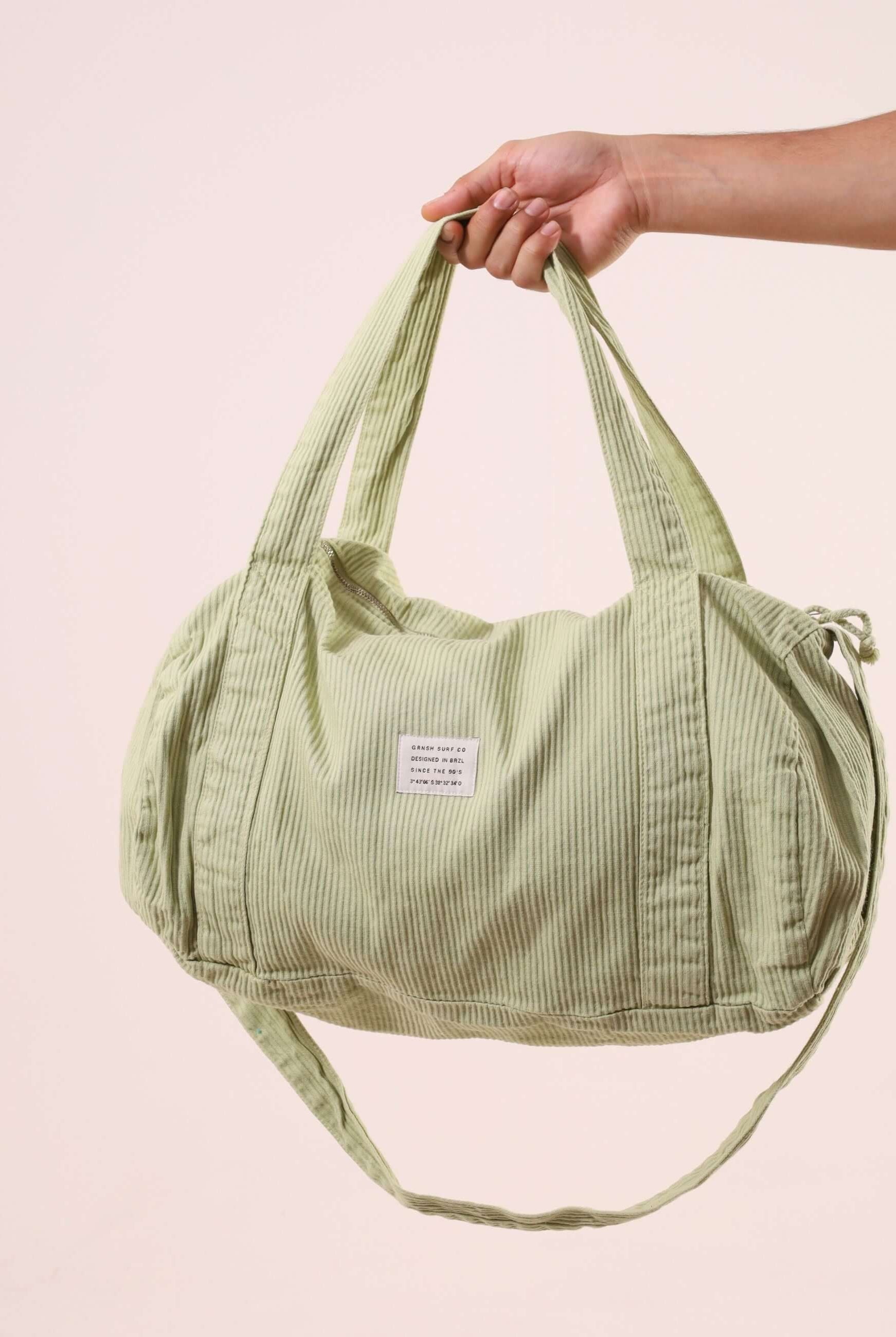 Hand holding a Sacola Duffel Bag - Verde Claro, showcasing its reinforced fabric handles and soft textured peletizada canvas in light green.