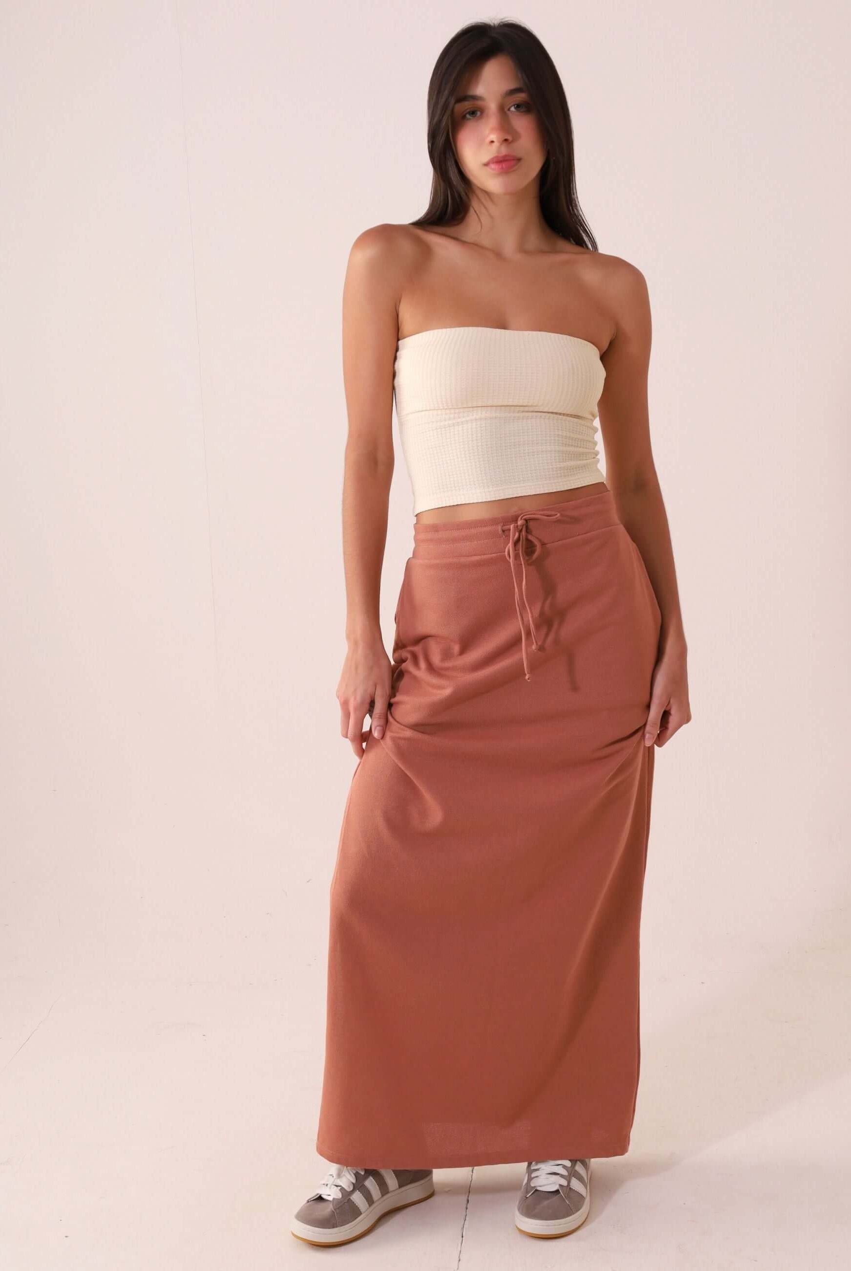 Mulher vestindo SAIA MIDI DÉA - CHOCOLATE da Greenish; saia reta e canelada, cós elástico com cadarço, fenda lateral direita.