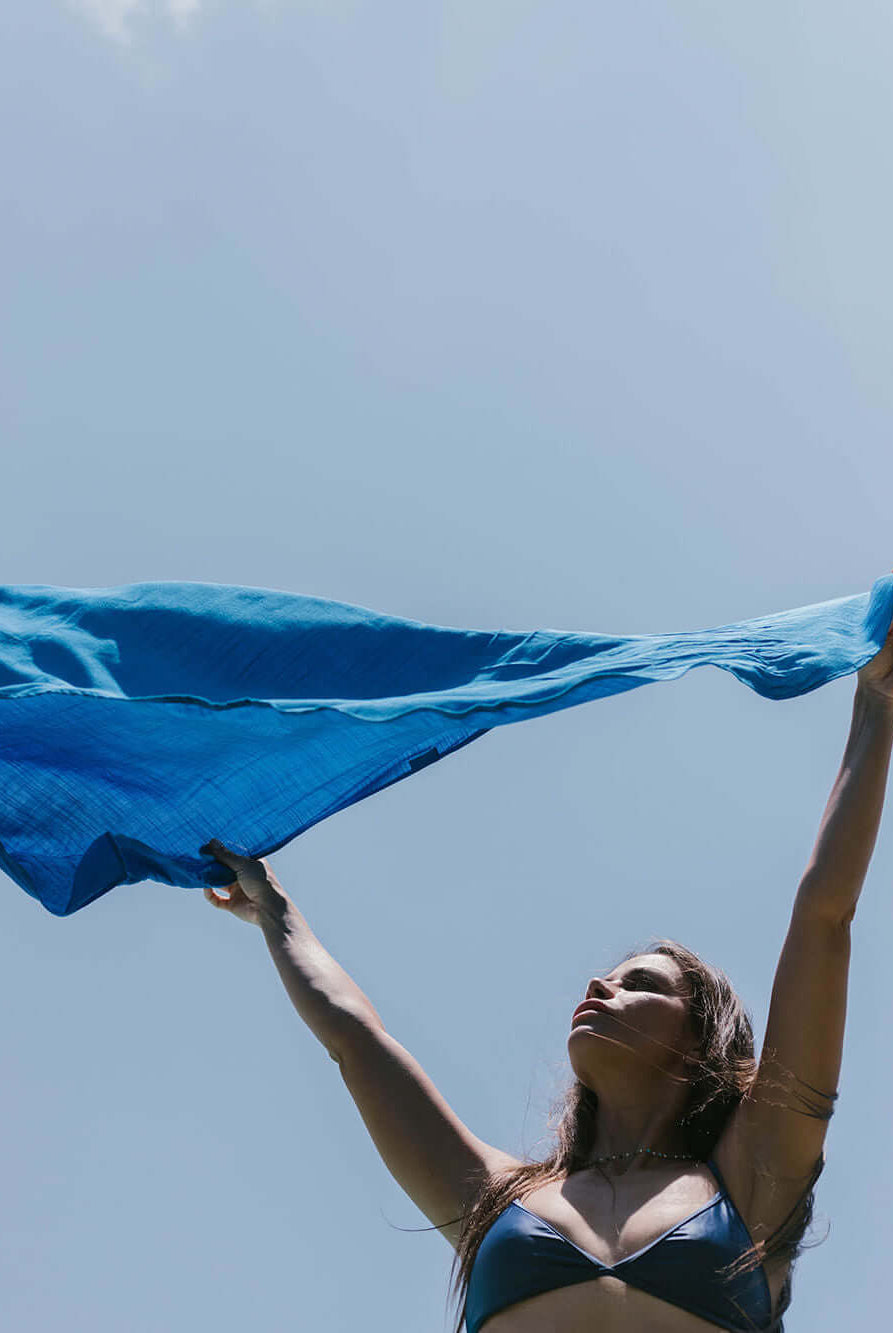 Mulher segurando sarong azul Praia Lana da marca Greenish ao ar livre, sob o céu claro.