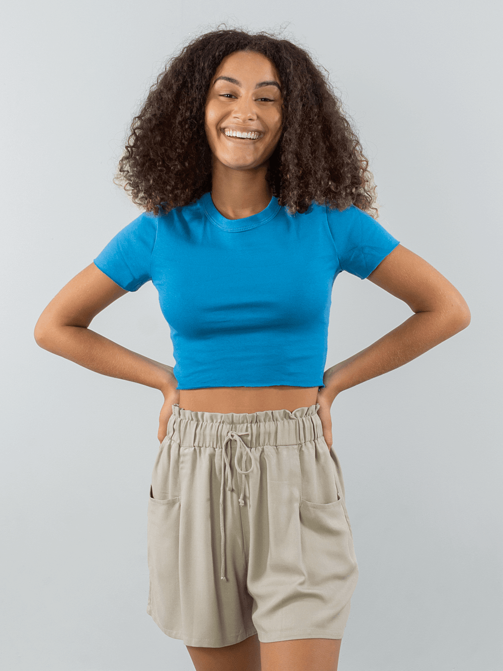 Modelo sorrindo usando blusa azul e short bege, da marca Greenish, destacando estilo casual e moderno.