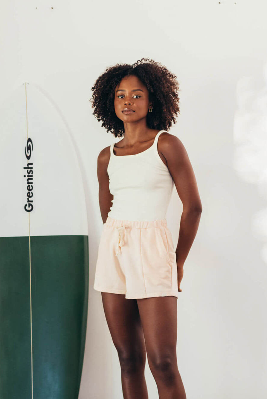 Modelo vestindo SHORT MOLETOM CLAY LARANJA da marca Greenish, com surfboard ao fundo, em ambiente iluminado.