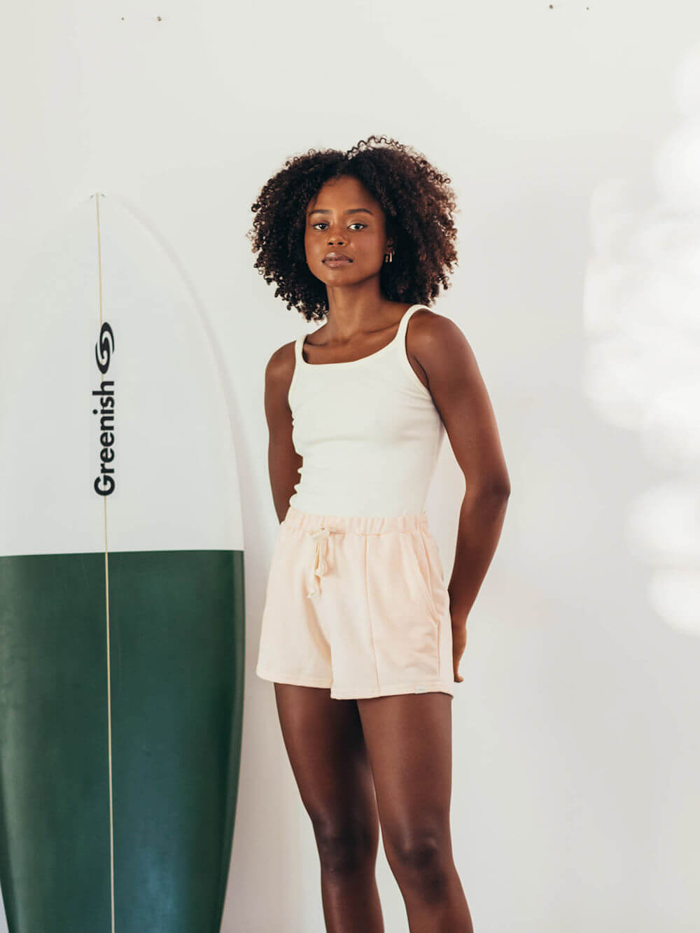 Modelo vestindo SHORT MOLETOM CLAY LARANJA da marca Greenish, com surfboard ao fundo, em ambiente iluminado.