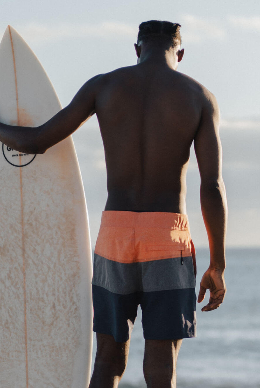Homem de costas usando boardshorts laranja e azul da Greenish, segurando uma prancha de surfe na praia.