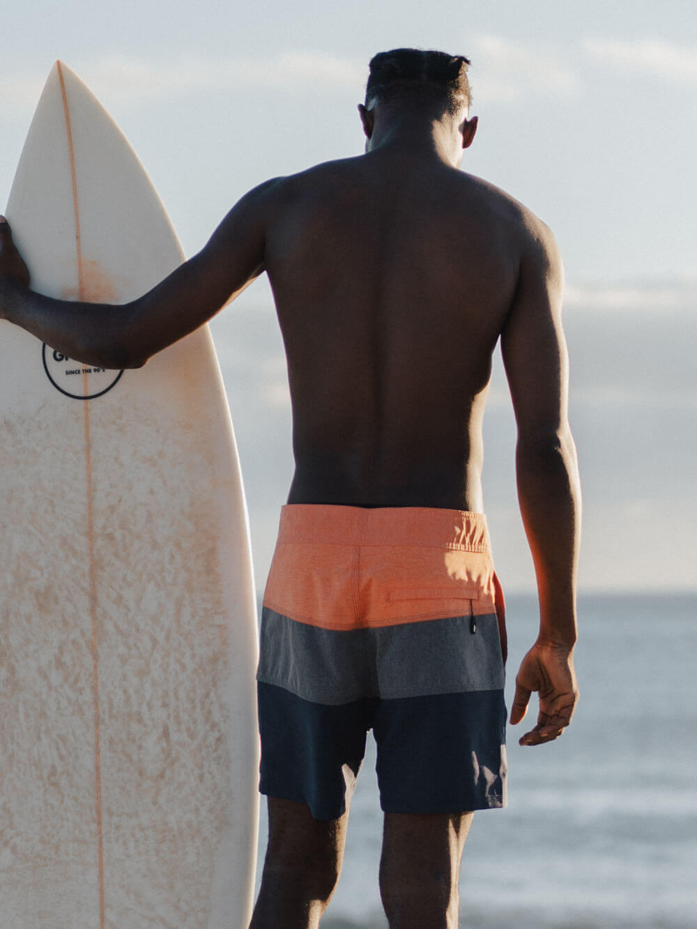 Homem de costas usando boardshorts laranja e azul da Greenish, segurando uma prancha de surfe na praia.