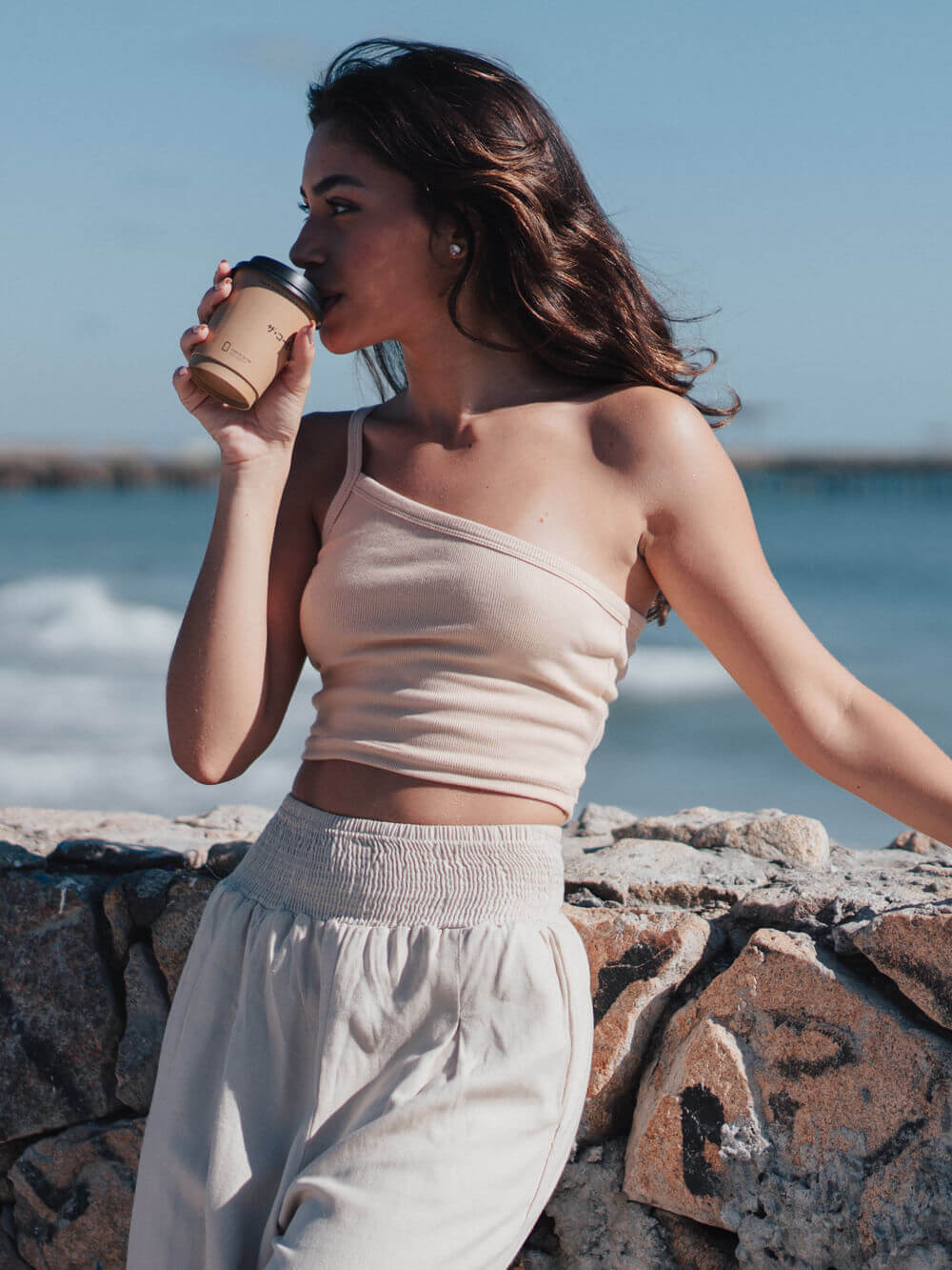 Top Zoe bege da Greenish, mulher tomando café à beira-mar, look casual e elegante.