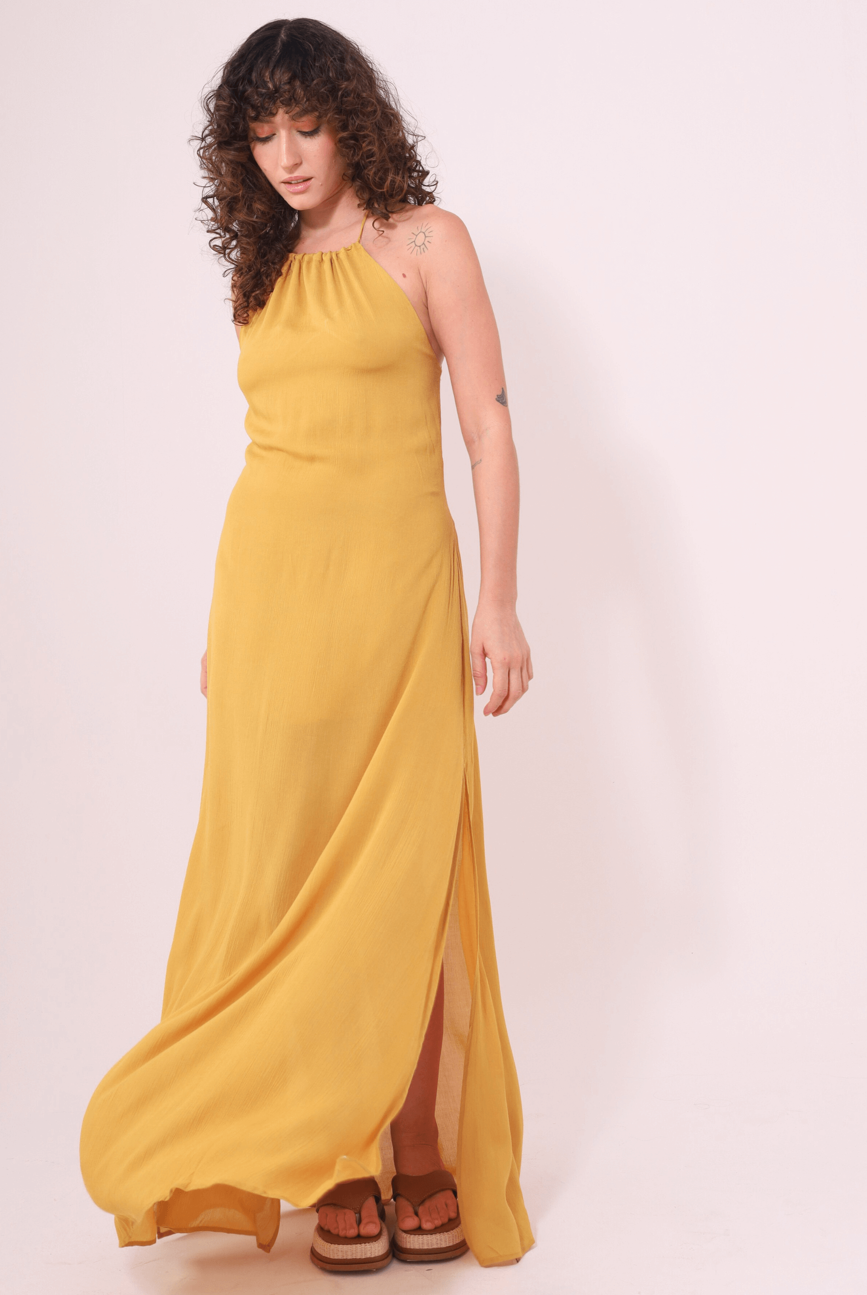 Mulher usando Vestido Longo Rachel Mostarda da marca Greenish, com alças finas e fendas laterais, ideal para dias quentes e elegantes.