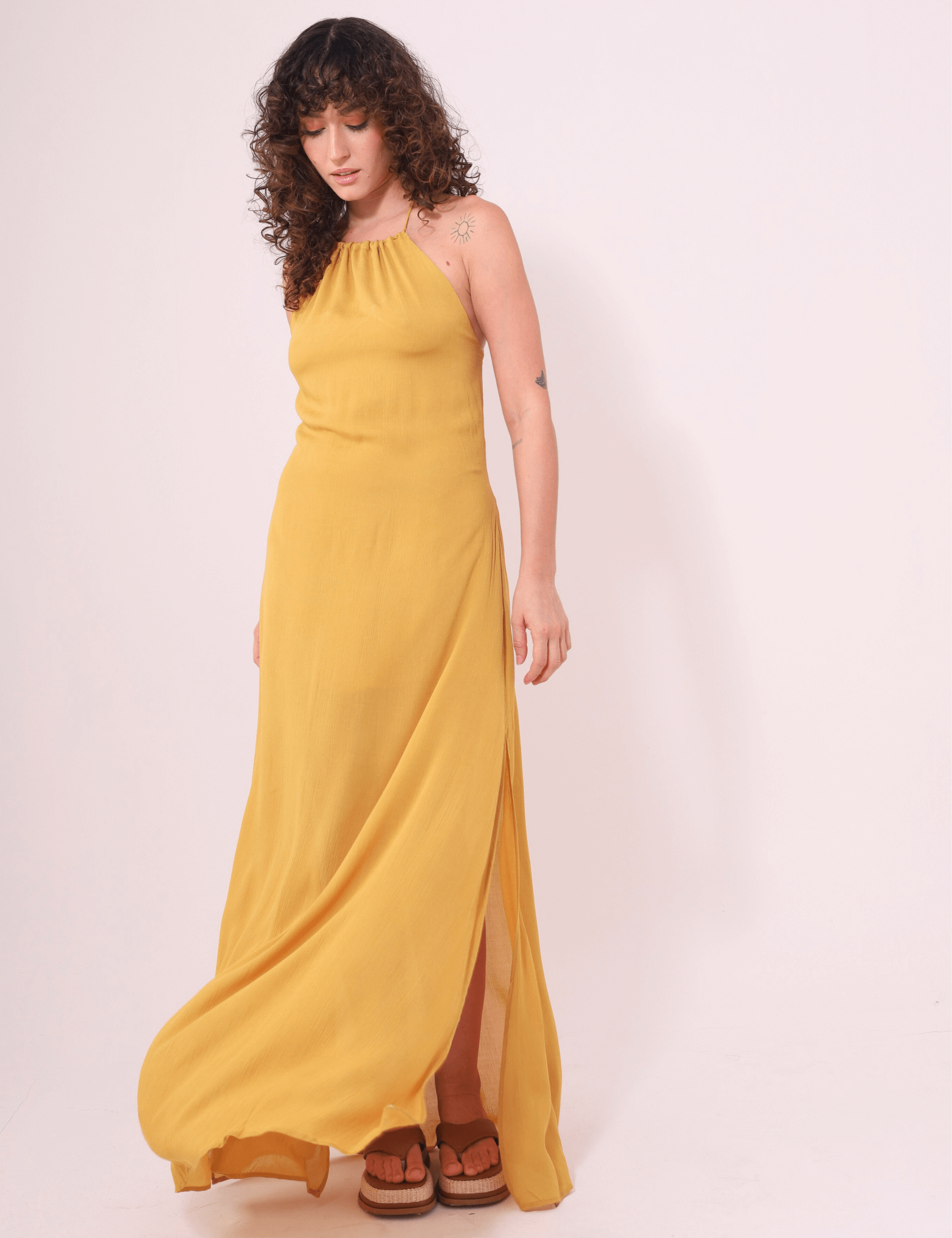Mulher usando Vestido Longo Rachel Mostarda da marca Greenish, com alças finas e fendas laterais, ideal para dias quentes e elegantes.