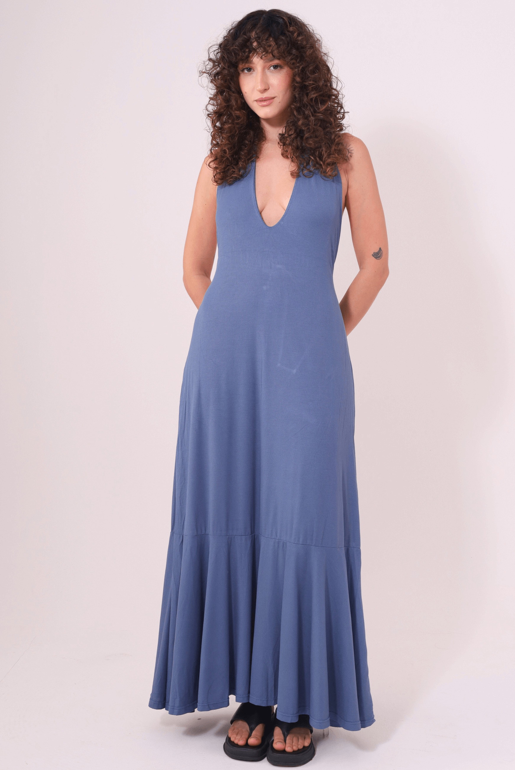 Mulher usando Vestido Longo Serena Azul Jeans com decote profundo e jabô na barra, da marca Greenish, ideal para dias de calor.