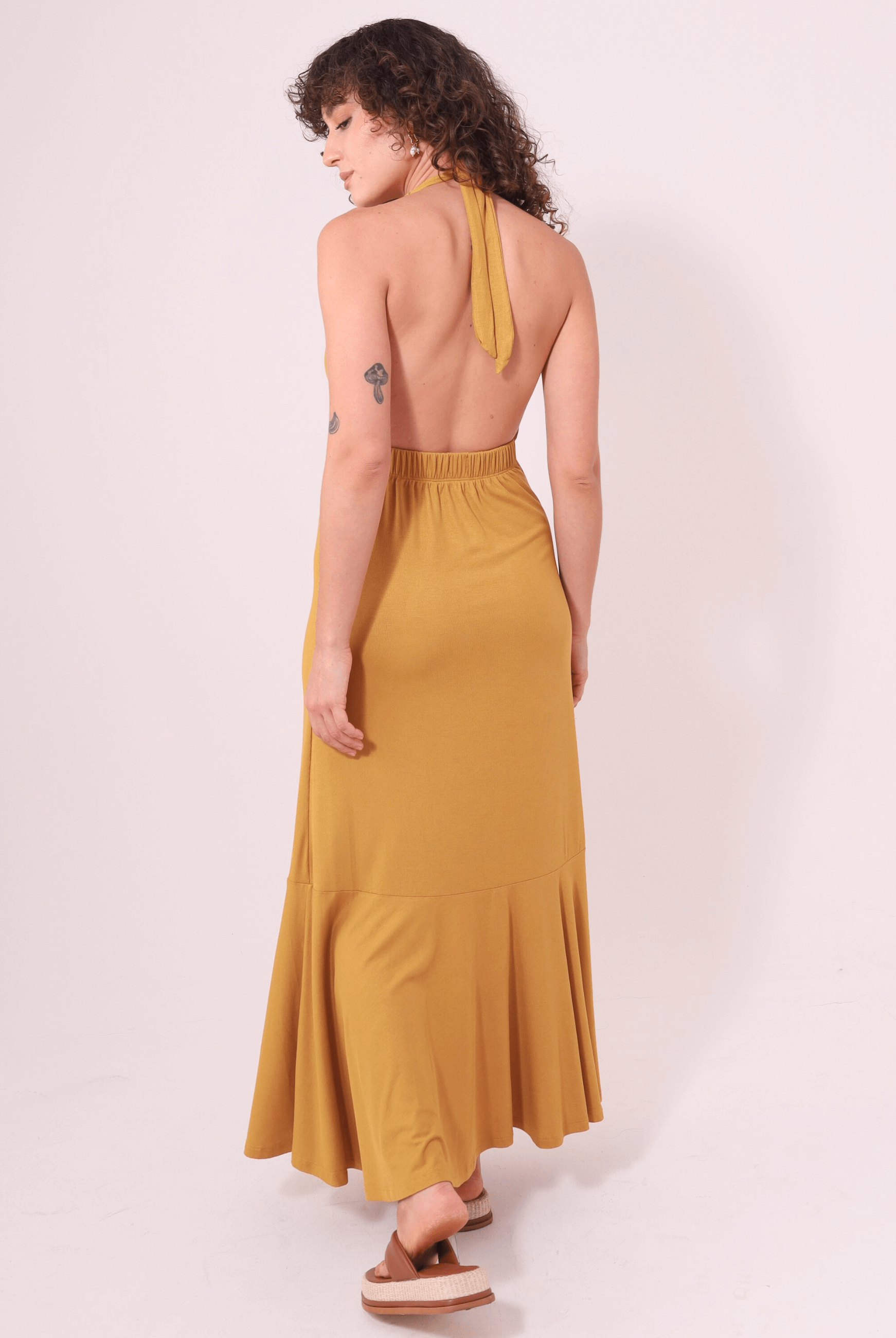 Mulher vestindo Vestido Longo Serena - Caqui da marca Greenish, com decote profundo e faixas no pescoço, em ambiente claro.