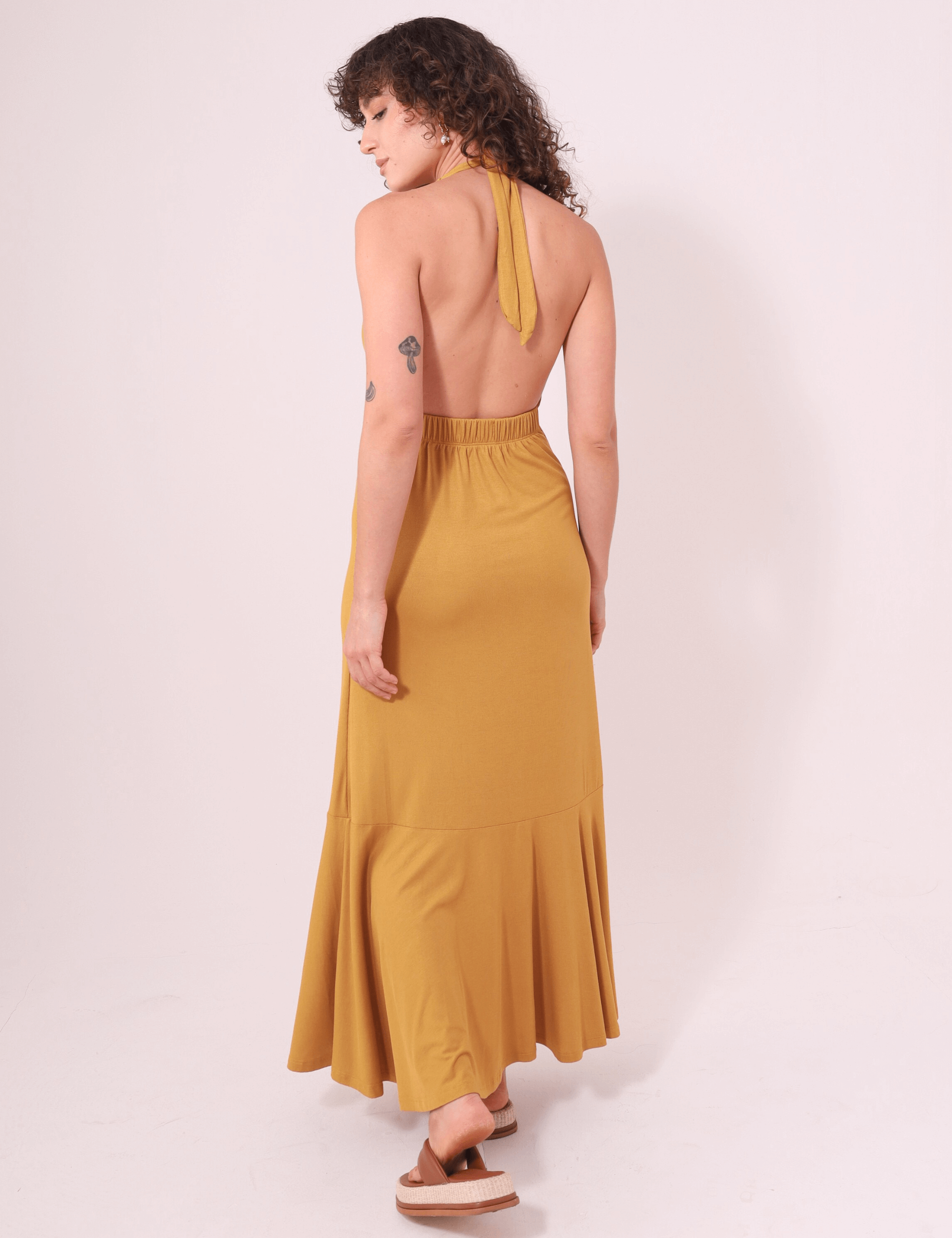 Mulher vestindo Vestido Longo Serena - Caqui da marca Greenish, com decote profundo e faixas no pescoço, em ambiente claro.