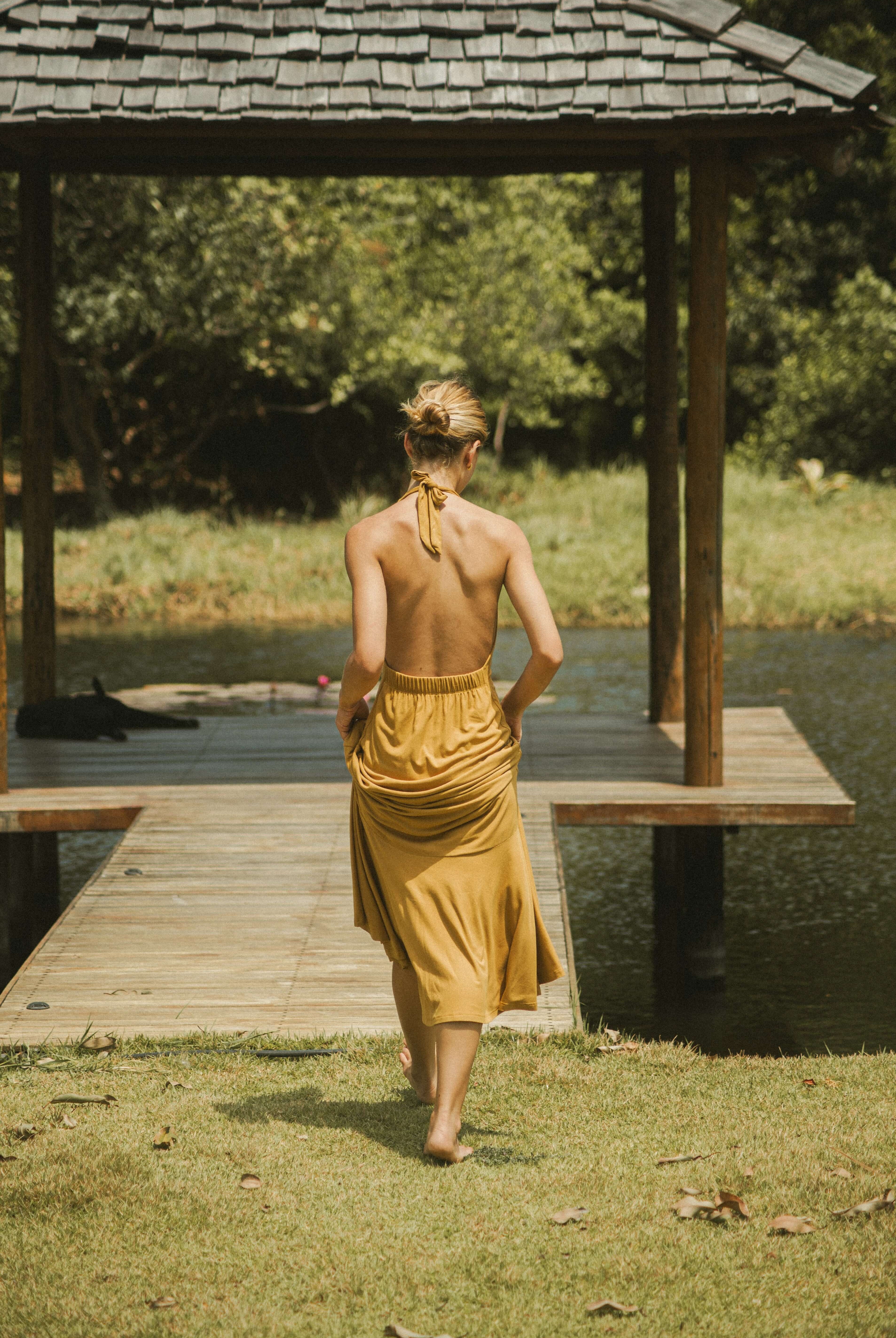 Mulher usando Vestido Longo Serena - Caqui da Greenish, caminhando em um píer com fundos naturais, destacando leveza e elegância.