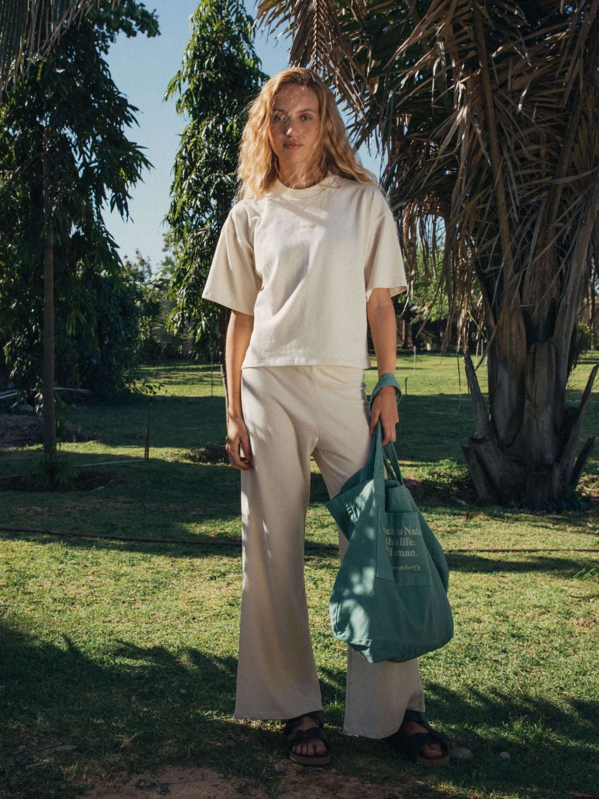 Modelo usando calça Gina bege da Greenish, em ambiente natural com palmeiras ao fundo.