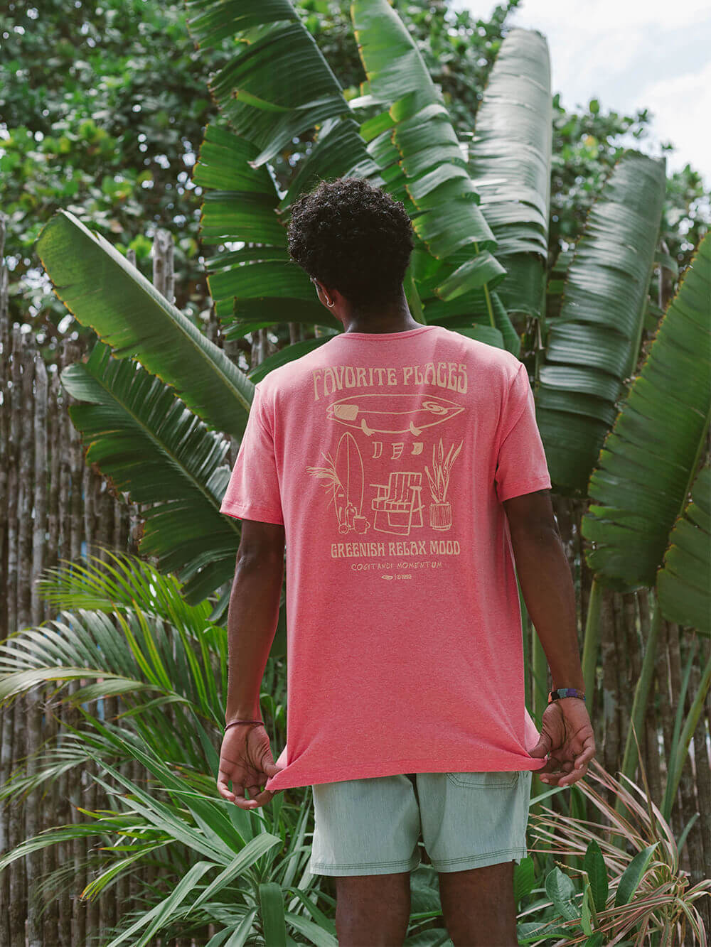 Model usando camiseta Greenish laranja com estampa, em ambiente tropical com palmeiras.