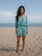 Mulher usando macaquinho Suzy turquesa da Greenish na praia, com decote em V e mangas longas.