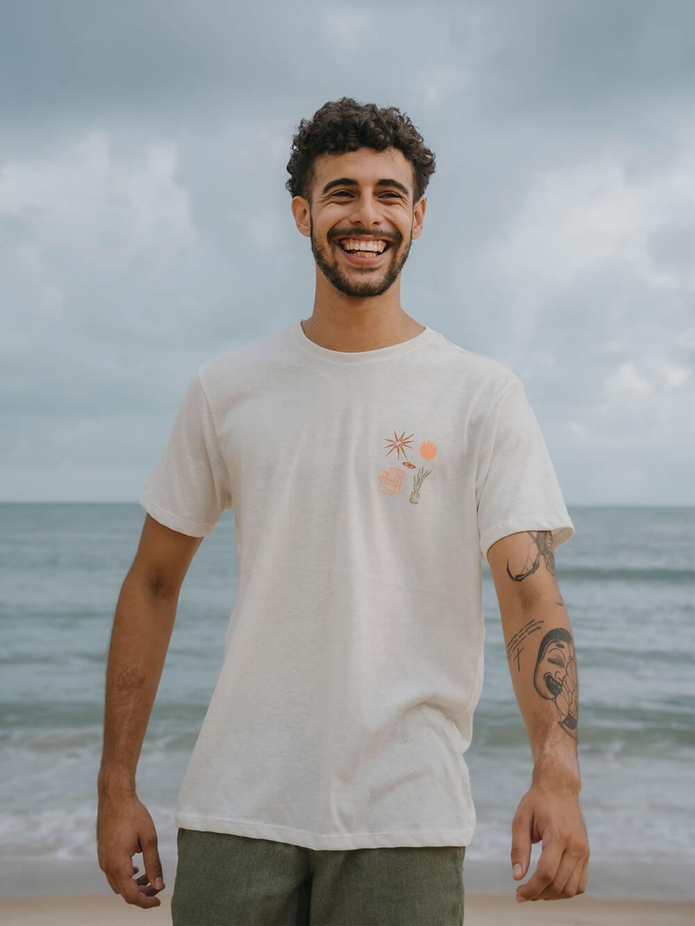 Camiseta Greenish Natural Elements bege, modelo sorrindo à beira-mar, destacando conforto e estilo simples.