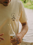 Homem usando uma camiseta Sandish Surf Co. enquanto segura uma bebida perto da água.