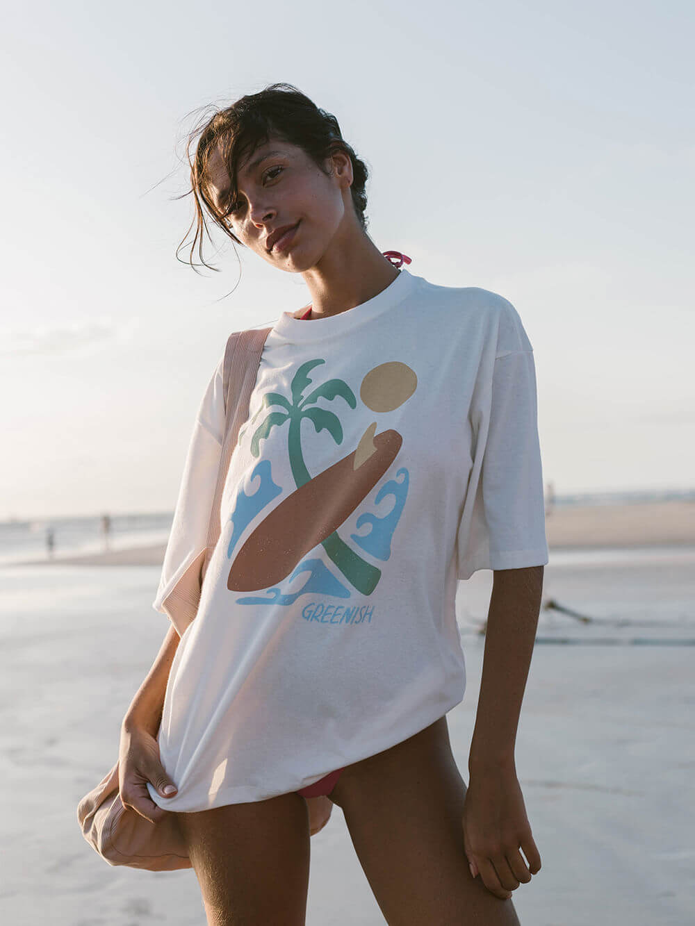 Modelo usando a camiseta oversized Sea Vibe da Greenish, na praia, com estampa tropical e estilo despojado.