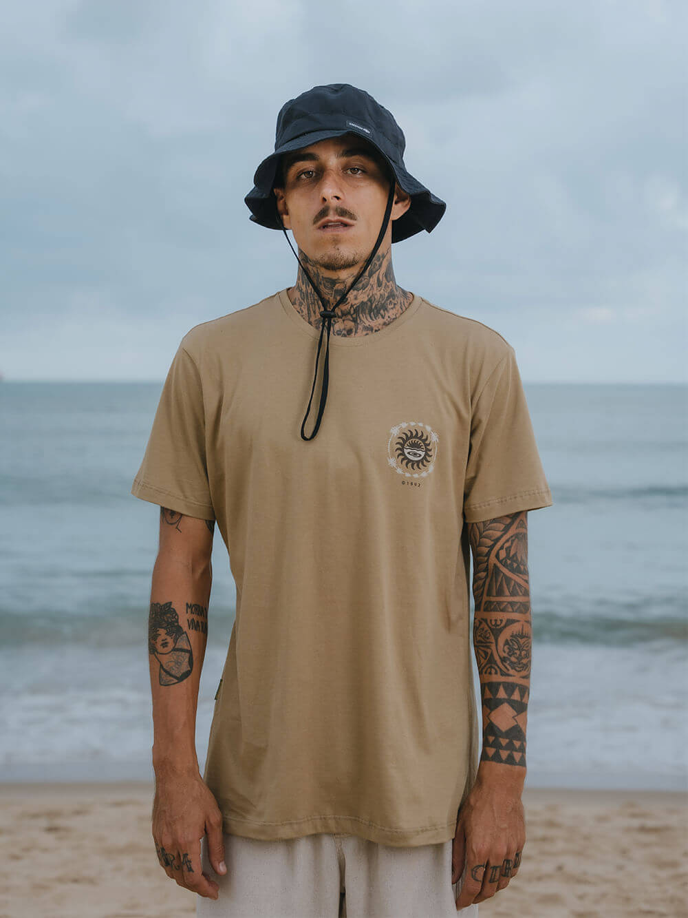 Modelo usando camiseta Elemental Suns caqui da Greenish na praia, com sol e estilo descontraído.