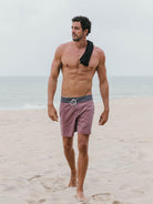 Homem com calções de banho bordô da Greenish, caminhando na praia. Estilo vintage e descontraído.