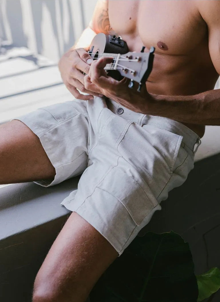 Homem tocando ukulele, vestido com shorts claros, sentado em uma superfície com luz natural.
