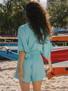 Mulher de costas usando macaquinho Suzy turquesa da Greenish em praia ensolarada.