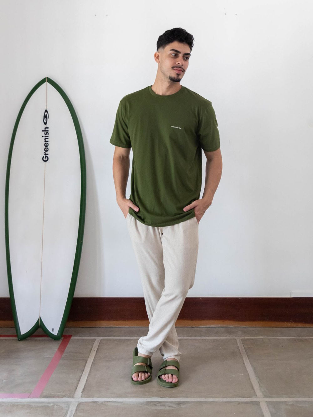 Homem usando camiseta verde escuro da Greenish ao lado de uma prancha, mostrando estilo casual e sustentável.