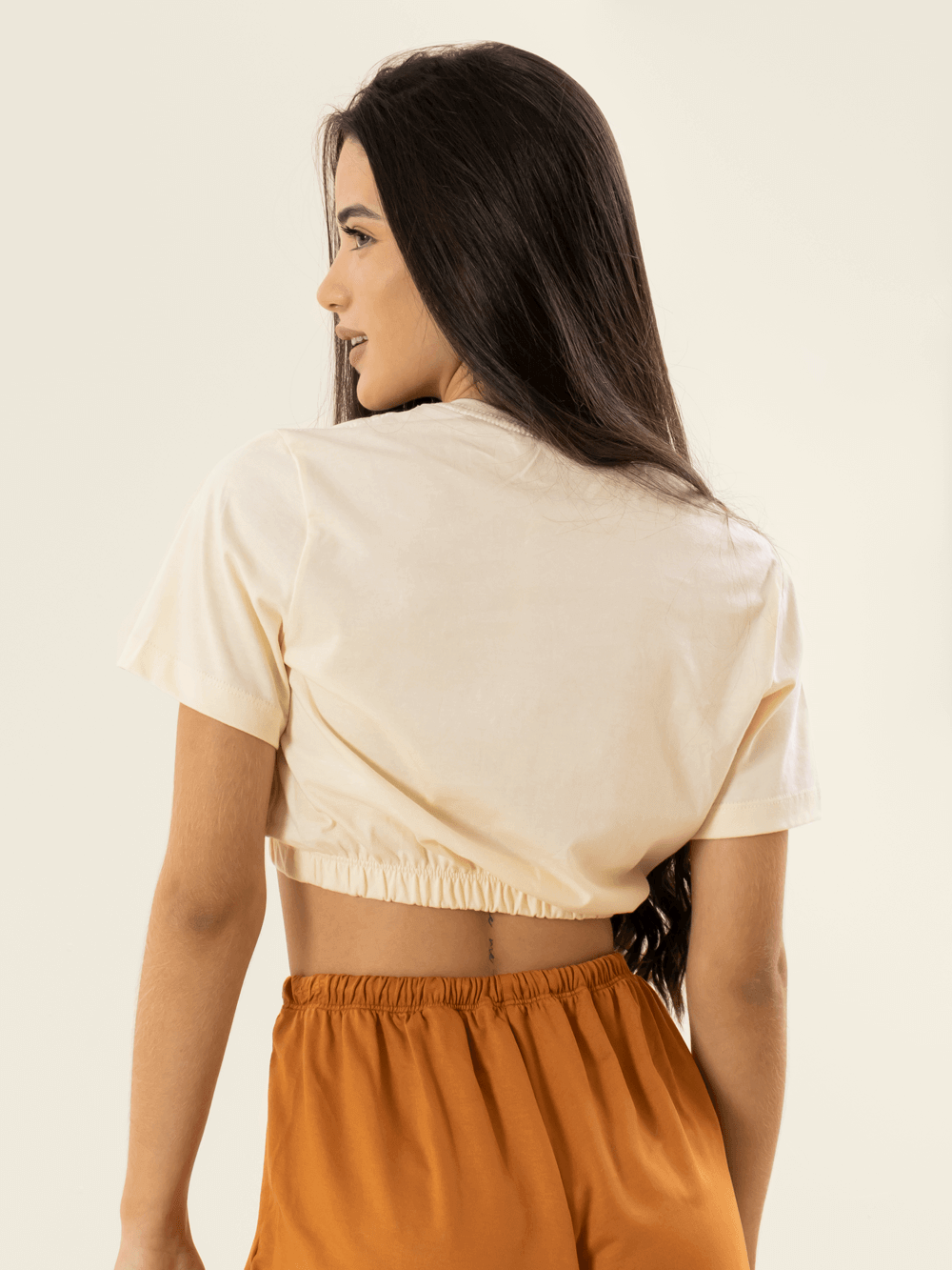 Mulher usando blusa cropped bege da marca Greenish, com detalhe franzido nas costas.