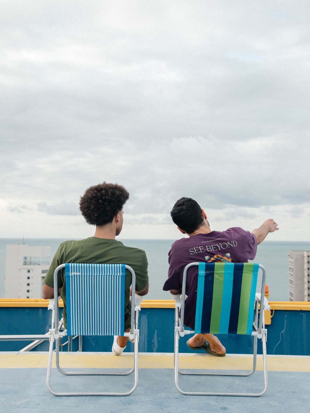 Dois homens relaxando em cadeiras de praia, observando o horizonte em um dia nublado.