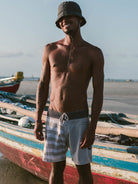 Modelo usando boardshort Yin Yang caqui da Greenish em praia ensolarada, com chapéu e barco ao fundo.