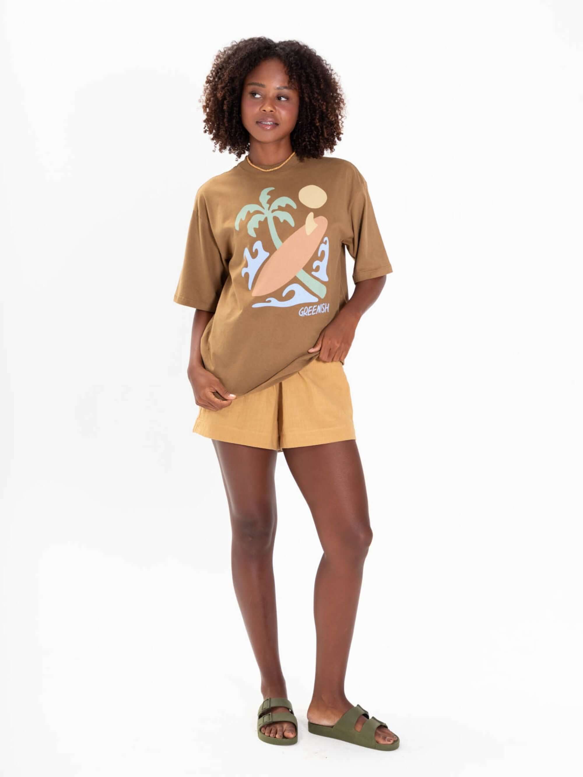 Modelo usando camiseta oversized caqui da Greenish com estampa de praia e palmeiras.