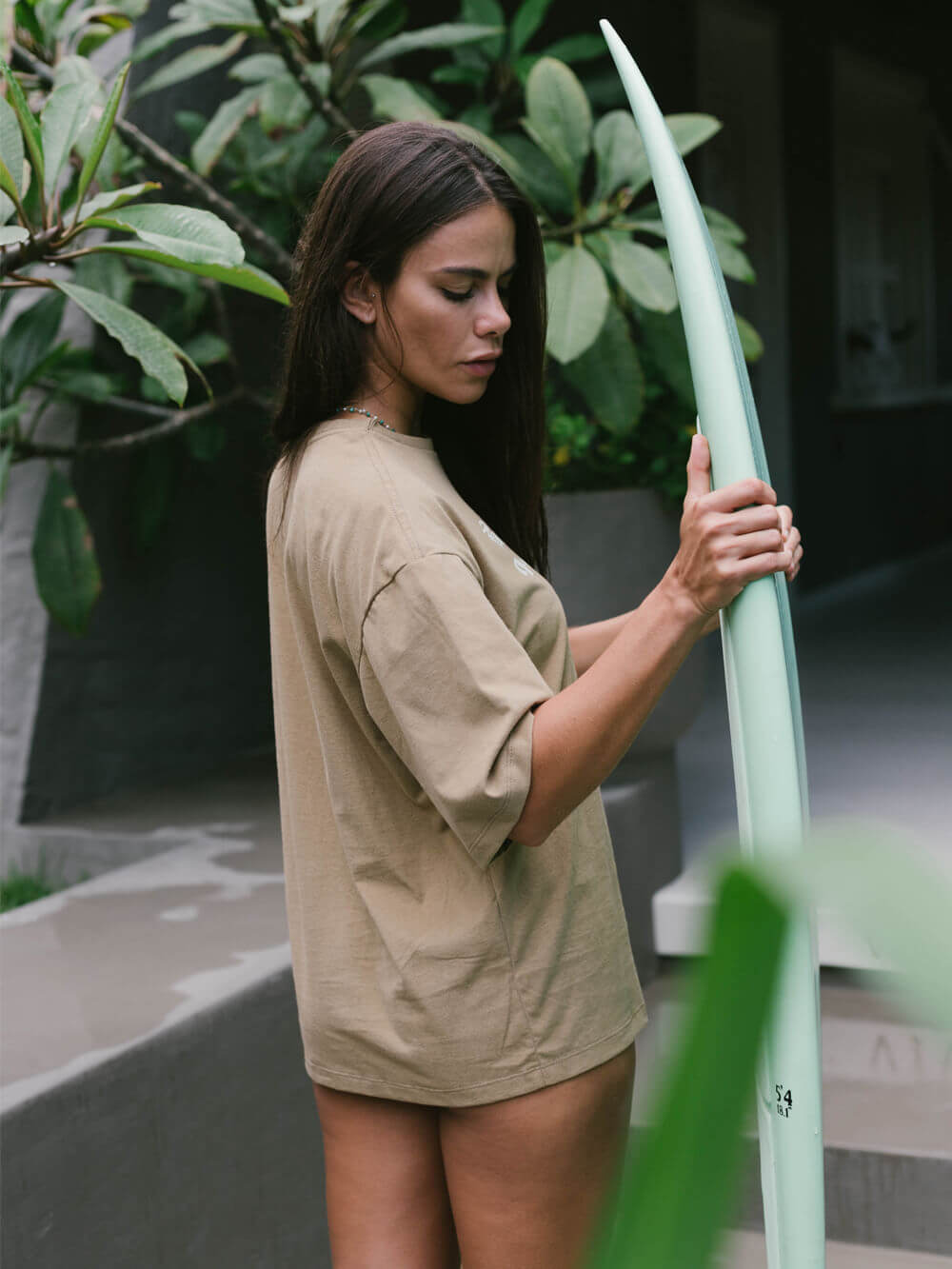 Modelo usando camiseta oversized Take Care Of Yourself da Greenish, em caqui, segurando prancha de surfe.