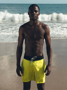 Homem usando o Boardwalk Sunny Viber amarelo da Greenish na praia, com ondas ao fundo, mostrando estilo e conforto.