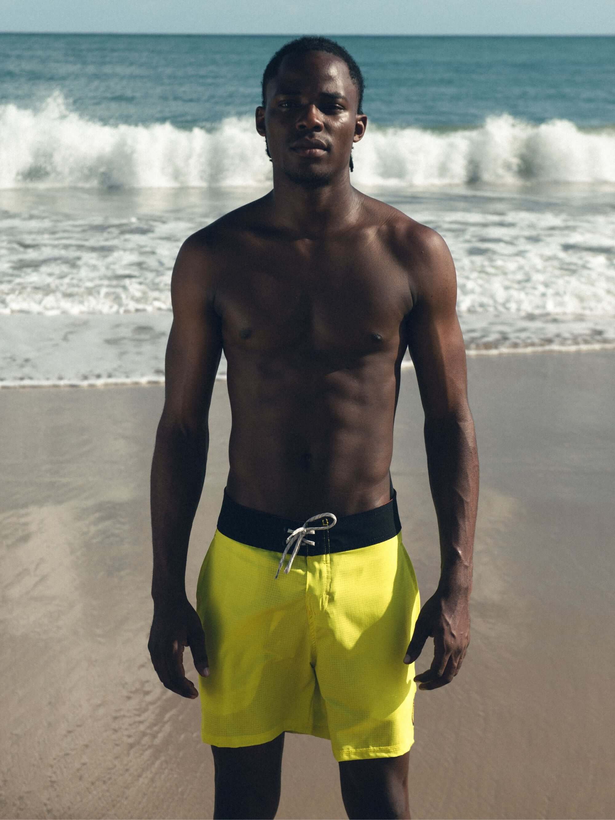 Homem usando o Boardwalk Sunny Viber amarelo da Greenish na praia, com ondas ao fundo, mostrando estilo e conforto.