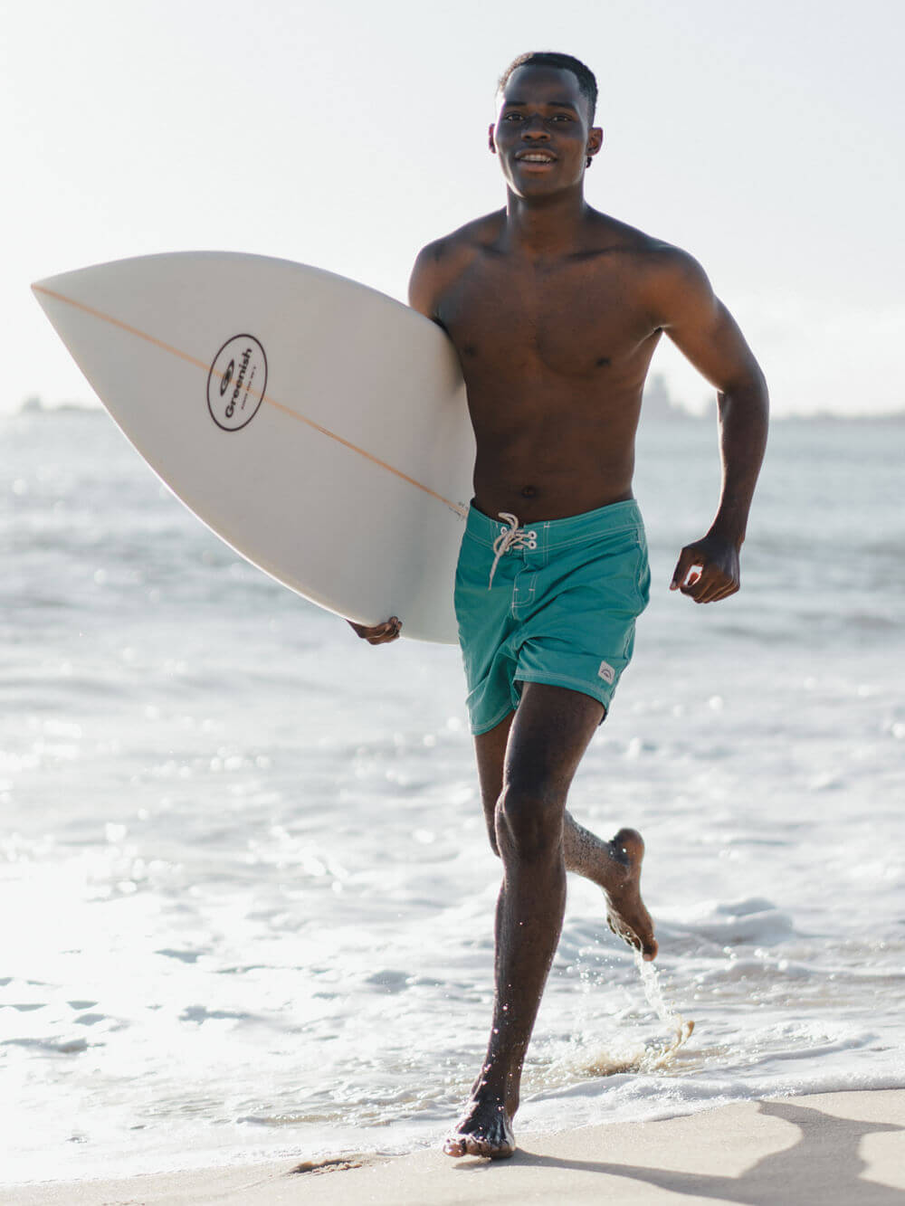Homem surfista correndo na praia, segurando uma prancha de surfe, usando bermuda azul-turquesa.