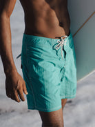 Modelo usando bermuda de banho verde-água ao lado de uma prancha de surf na praia.