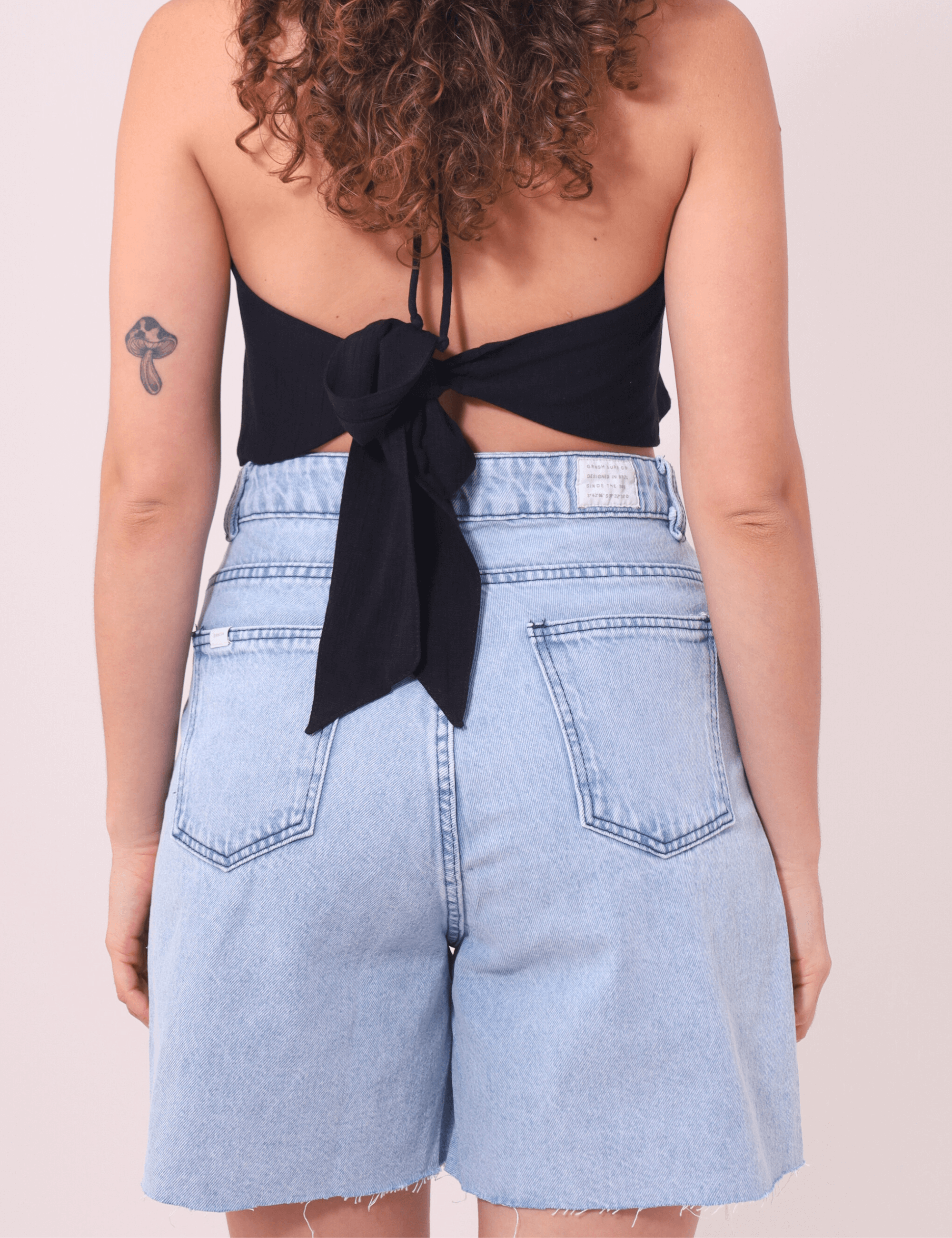 Bermuda Jeans Glicia Greenish cintura alta vintage azul, estilo e conforto para o verão. Modelo feminino de costas com top preto.