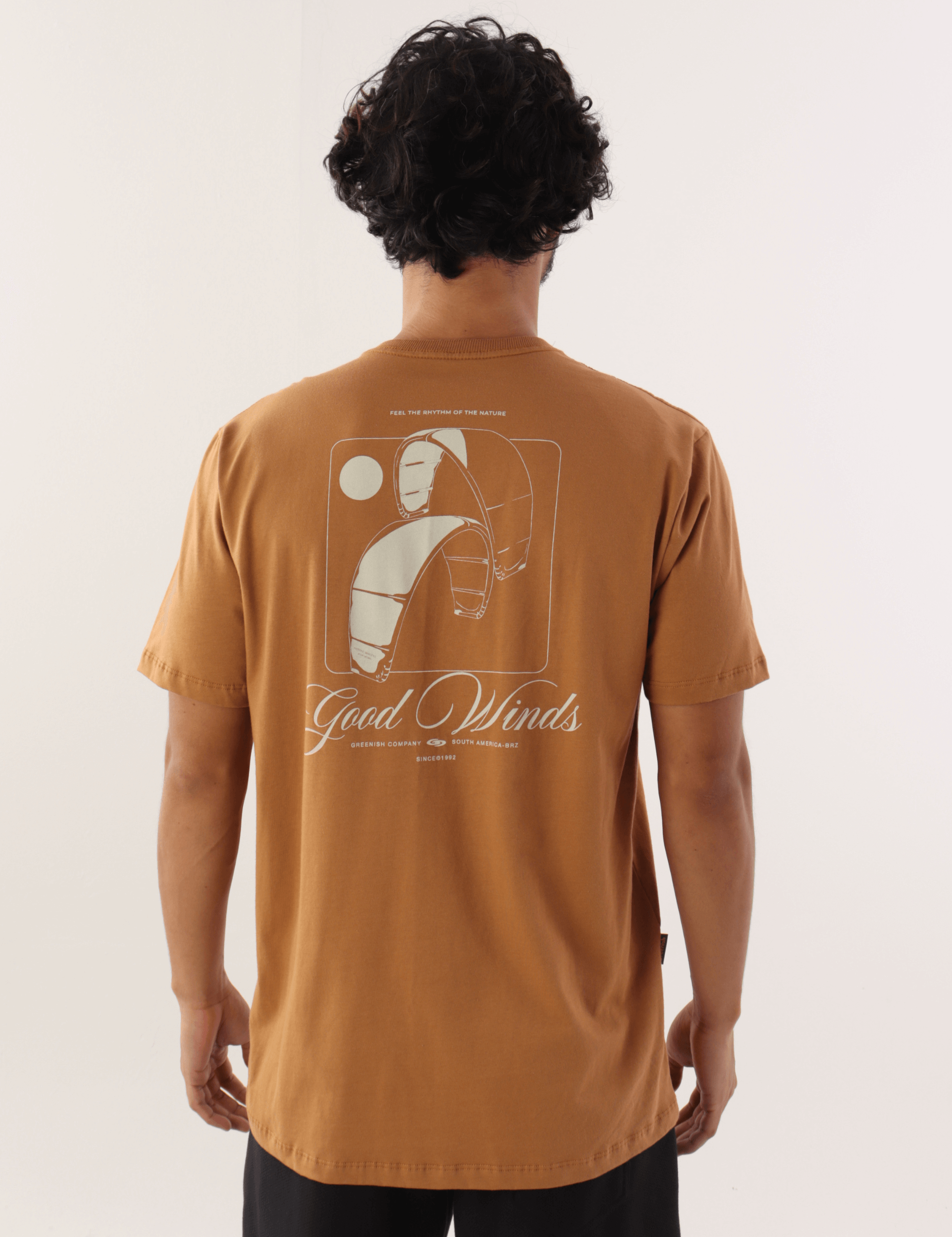 Homem usando camiseta marron da marca Greenish com estampa "Kites & Good Winds" nas costas, em estilo casual e esportivo.