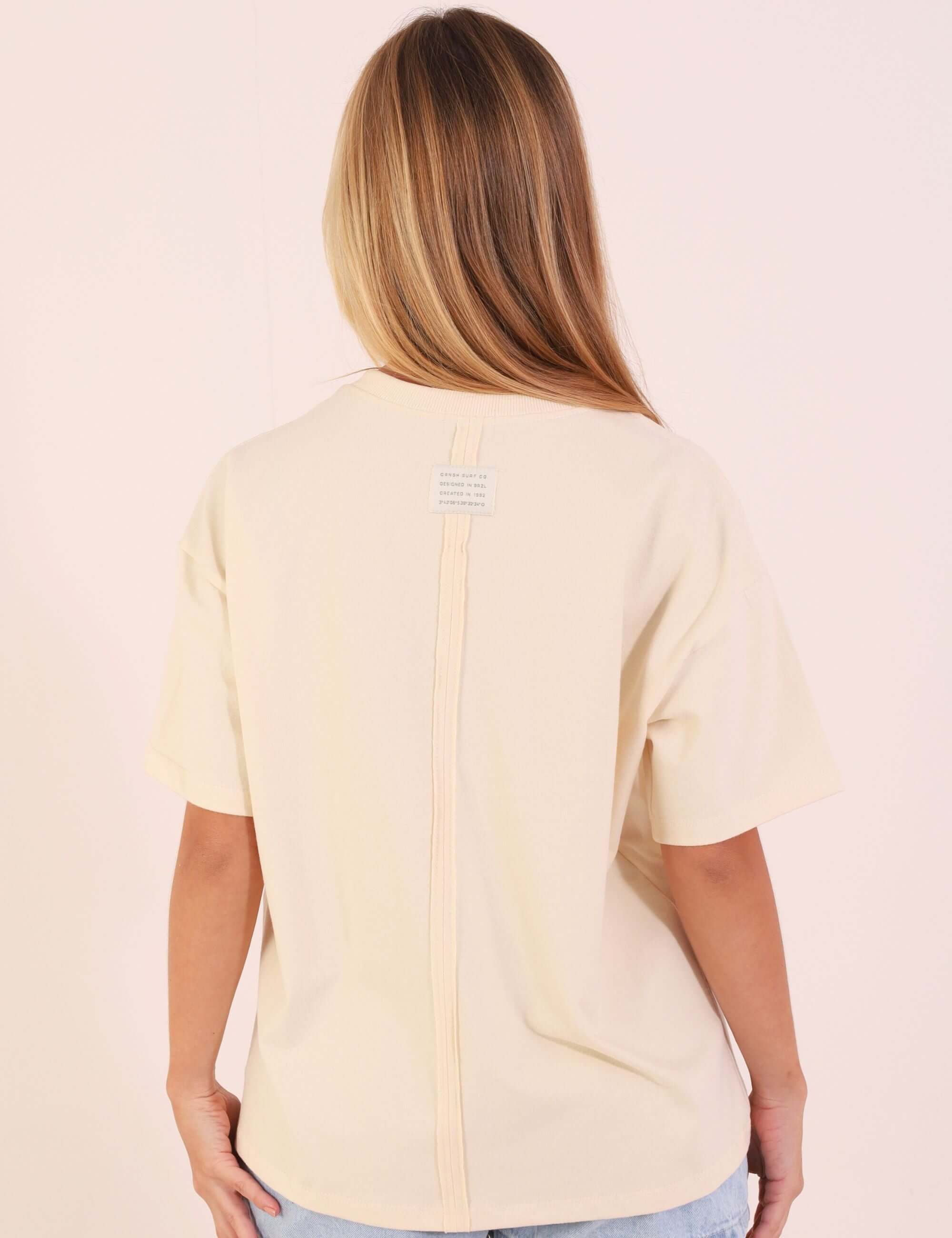 Modelo vestindo Camiseta Oversized Breeze - Bege em malha essencial com recorte moderno, vista de costas, ideal para looks casuais.