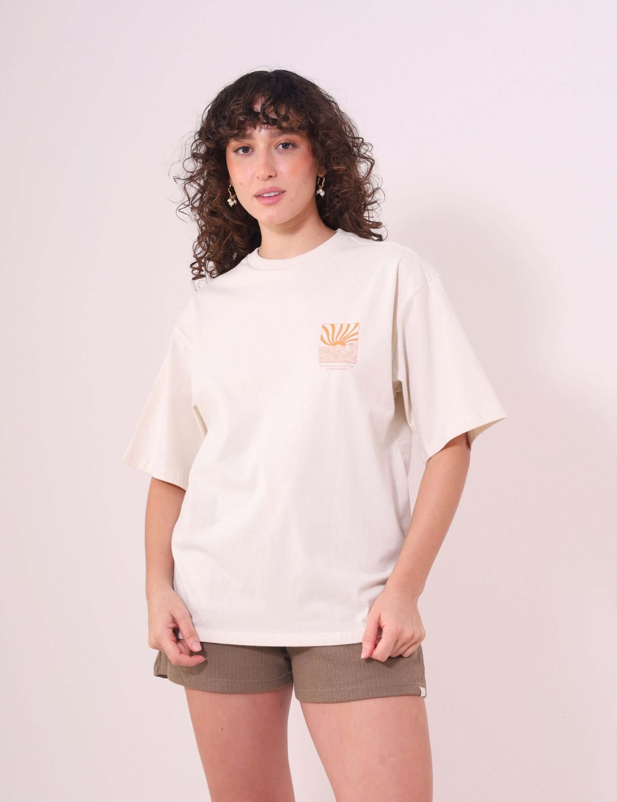 Mulher vestindo Camiseta Oversized Sunset Session da Greenish, cor natural, estampa de ondas e pôr do sol, feita de algodão macio.