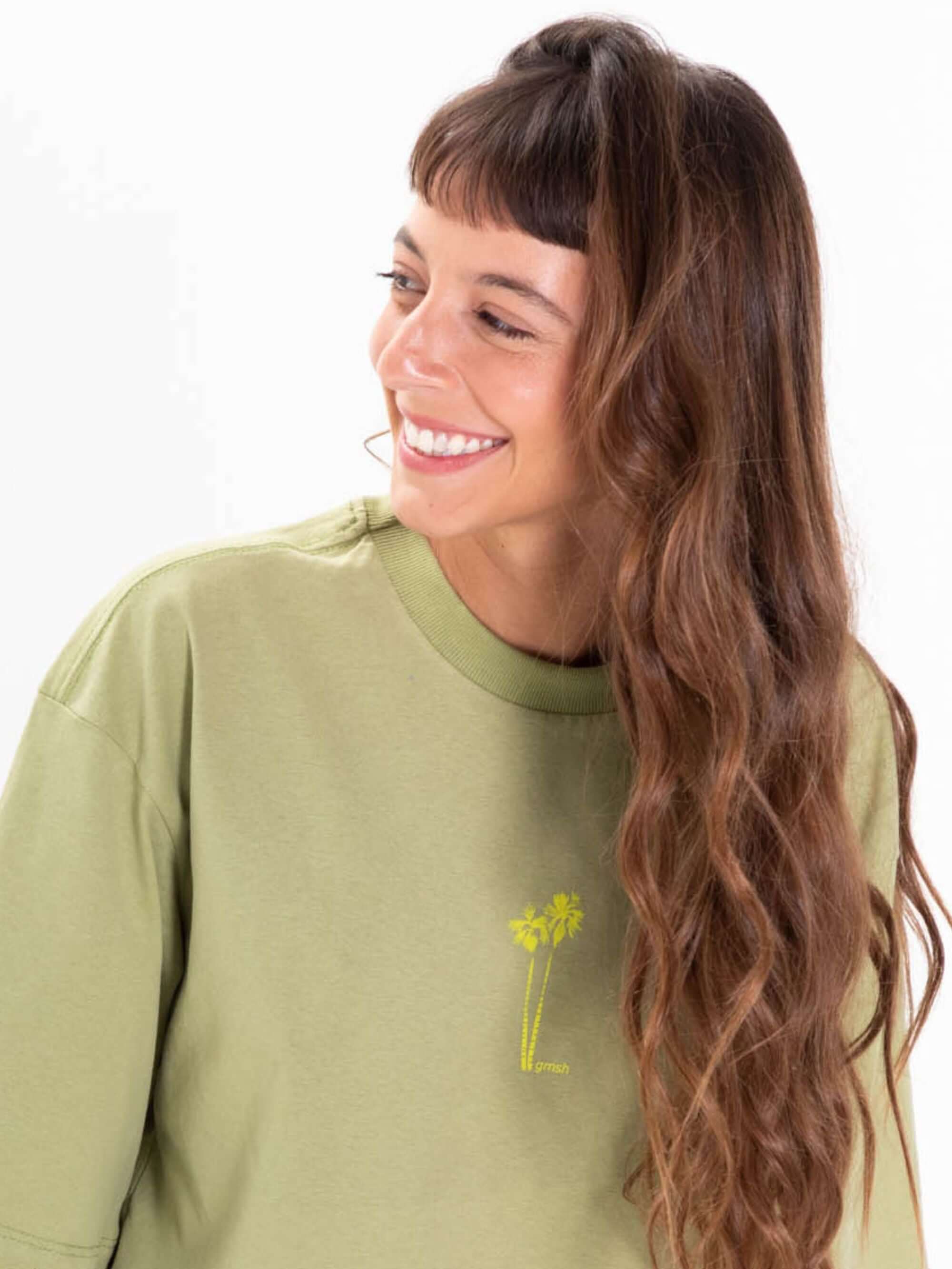Camiseta oversized verde com estampa de palmeiras da Greenish, modelo feminino sorrindo.