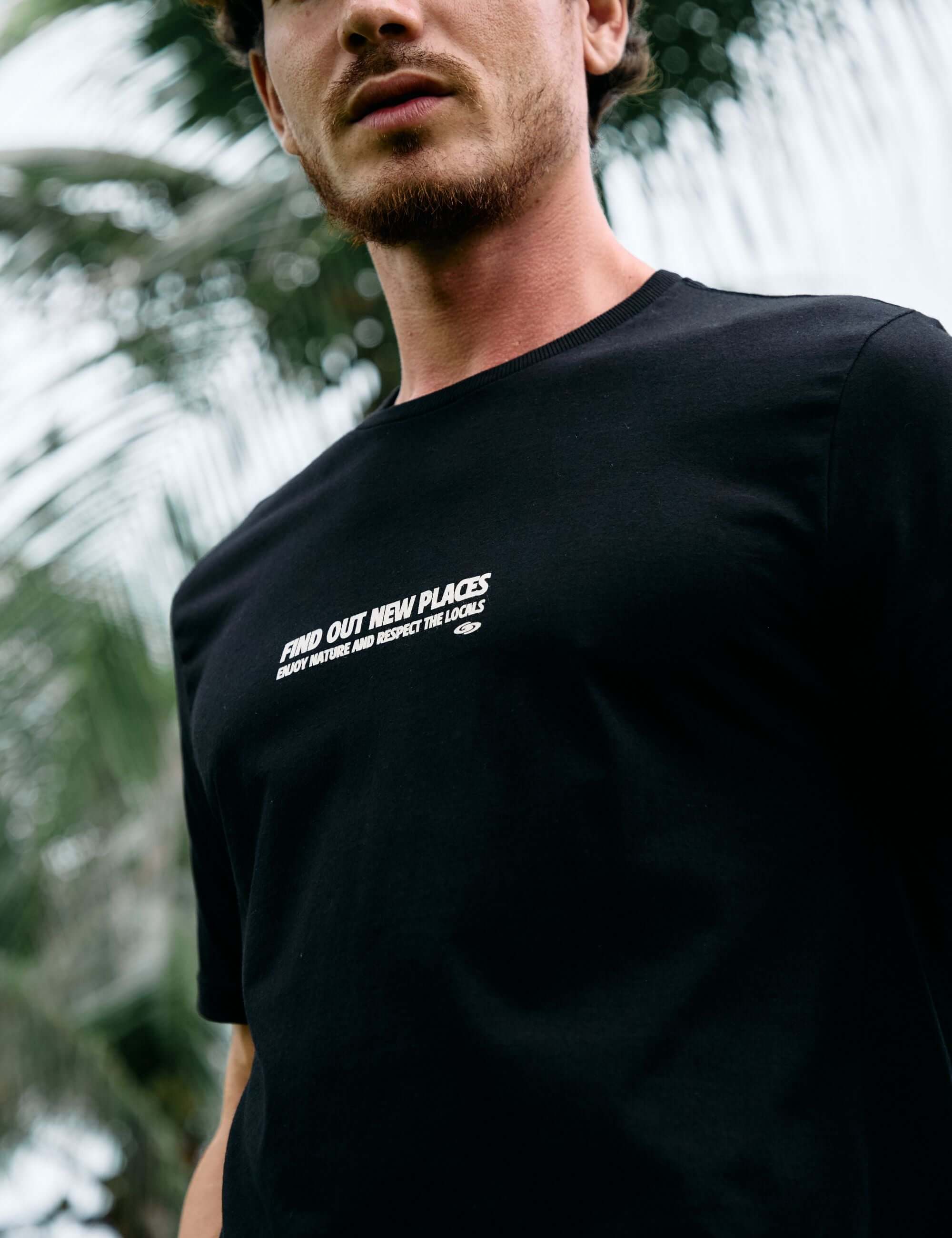 Homem vestindo camiseta preta Seagull Greenish com estampa, em meio à natureza tropical, representando liberdade e estilo.