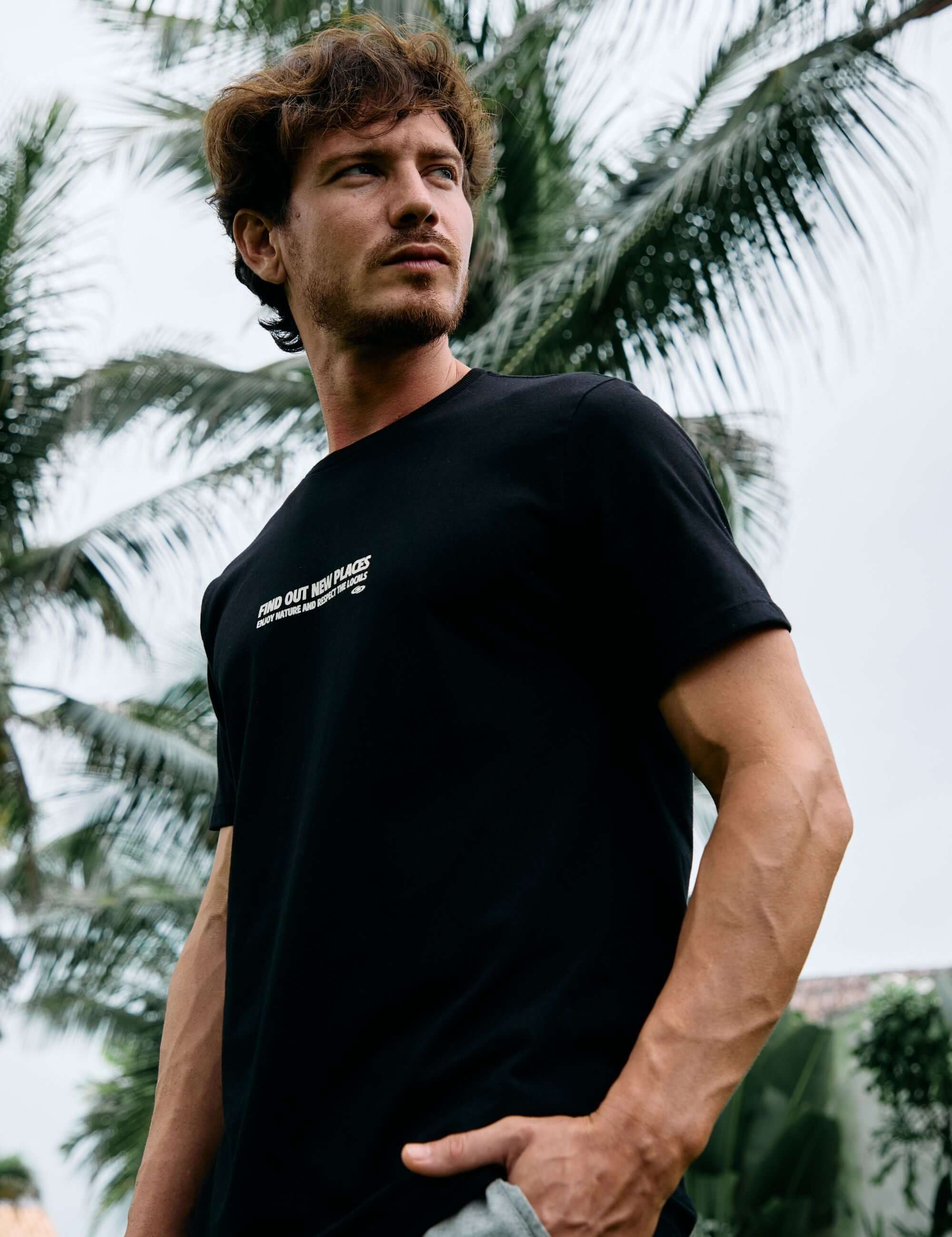 Homem usando camiseta preta Seagull Greenish ao ar livre com palmeiras ao fundo, evocando liberdade e natureza. 100% algodão.