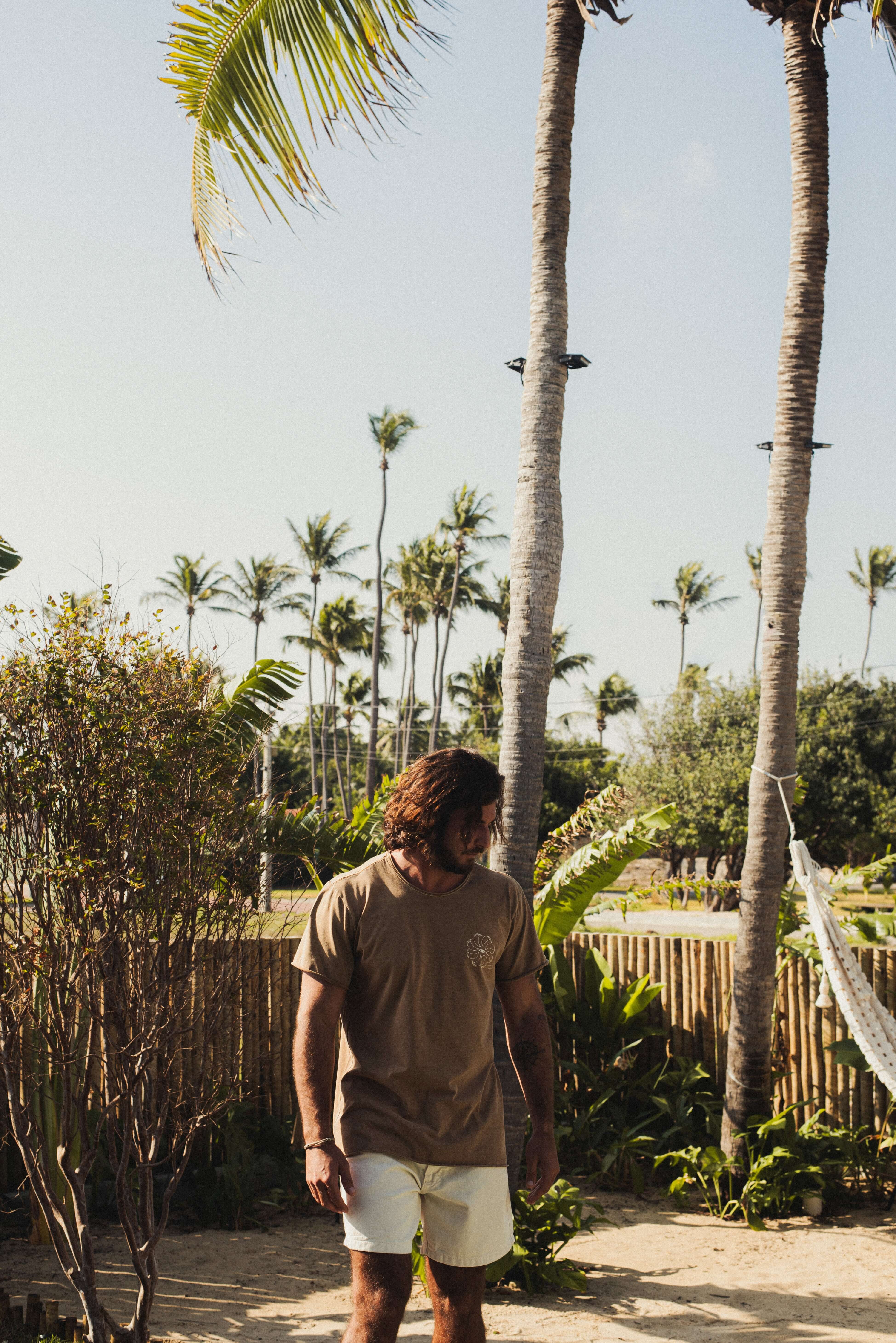 Homem vestindo Camiseta Stone Feel The Winds - Marrom da Greenish, em um cenário tropical. Toque macio e visual rústico. 100% algodão.